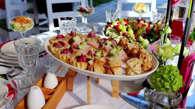 Elegant oval plate displaying gourmet appetizers, pastries, and ham on wooden support, surrounded by decorative dining elements creating refined table setting
