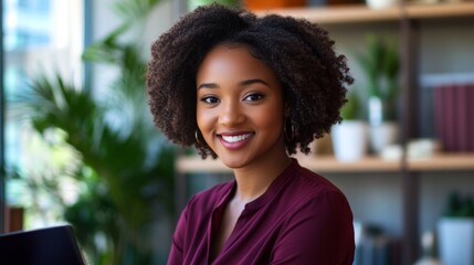 A woman with curly hair is smiling and sitting in front of a laptop. She is wearing a red shirt and has a gold hoop earring