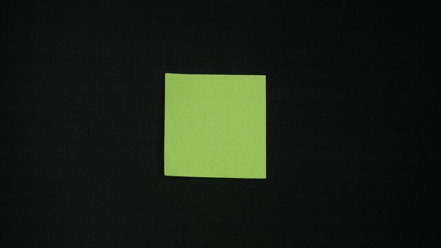 A woman's hand is sticking a yellow sticker onto a black wall. A young woman is sticking a note on a black wall