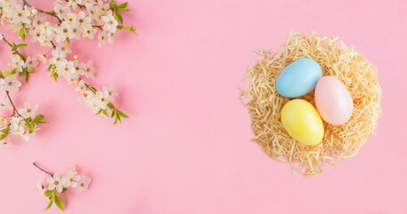 Colored easter eggs in a nest of straw and white flowering tree branches on the pink background. Top view. Copy space.