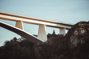 Modern concrete bridge with large angular supports spanning a lush green hillside with rocky cliffs...