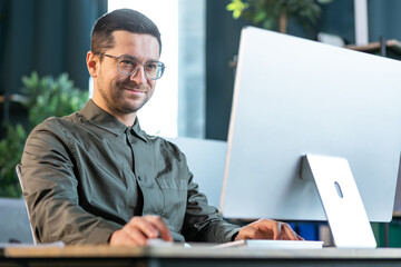 Smiling creative business man typing on desktop computer in office. Happy man wearing eyeglasses working on his computer at modern office space