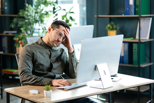 Portrait of sad bored business man sitting at desk using pc, leaning head on hand looking at screen. Upset stressed guy suffering job problems, reading bad negative news at office, free copy space