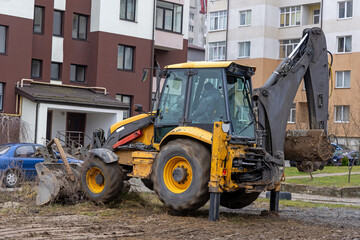 Backhoe loader working near residential buildings. A yellow backhoe loader actively digging in a muddy area near residential apartment buildings.