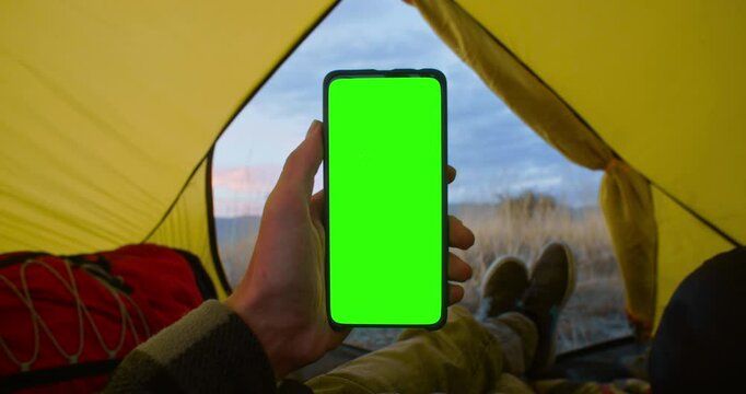 Person wearing a jacket is inside a cozy tent holding mobile phone and not touching touch screen. Use green screen for copy space closeup. Chroma key mock-up on smartphone in hand.