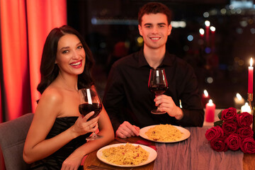 Portrait of young European couple celebrating St. Valentine's Day at restaurant, drinking red wine and smiling at camera, focus on woman