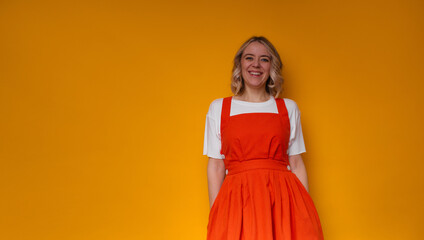 A cheerful woman stands against a vibrant yellow background, wearing a stylish orange dress over a...