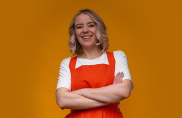 Confident woman in an orange dress and white shirt, arms crossed, smiling against a vibrant yellow...