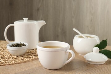 Refreshing green tea in cup, sugar bowl, leaves and teapot on wooden table