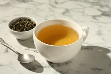 Refreshing green tea in cup, dry leaves and spoon on white marble table, closeup