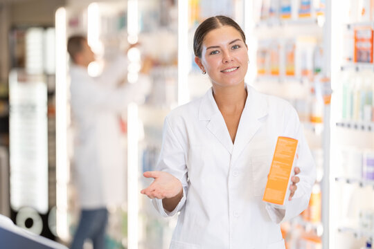 Smiling young woman chemist demonstrating medicament in box in pharmacy store