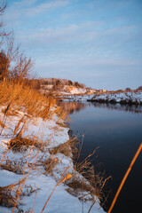 A picturesque snowy shoreline of a serene river, with a fishing rod prominently placed in the foreground, creating a charming winter scene that captures the essence of natures beauty