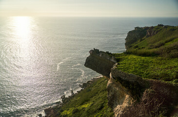 The breathtaking view showcases a stunning cliff adorned with lush greenery and a sparkling ocean during sunset