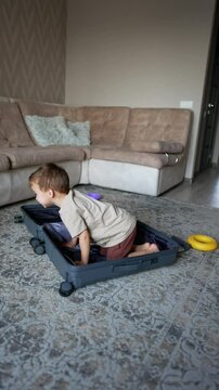 Cute Caucasian boy is hiding in the big suitcase. Kid stands up and goes to pick up his clothes scattered by the room. Vertical video.
