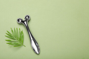 Metal face roller and palm leaf on green background, flat lay. Space for text