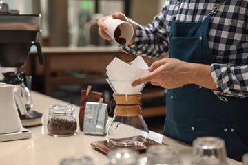 Barista pouring ground coffee into glass coffeemaker with paper filter at table in cafe, closeup
