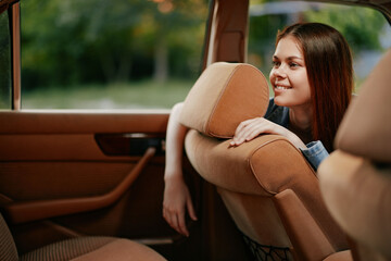 Young woman in casual attire smiling and leaning out of a car window, surrounded by greenery,...