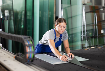 Skilled young female worker carefully cutting sheet of glass on table using manual glass cutter in industrial factory setting..