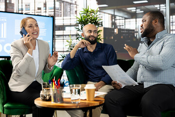 Happy woman talking on phone next to coworkers looking over revenue figures on documents in office. Cheerful worker in telephone call next to colleagues comparing company statistical numbers
