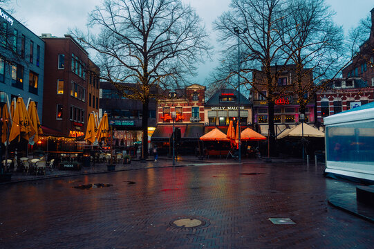 Evening view of a lively city square in Enschede with lit cafes, restaurants, and trees adorned with fairy lights. Reflective wet pavement. Enschede, Netherlands. January 6 2025