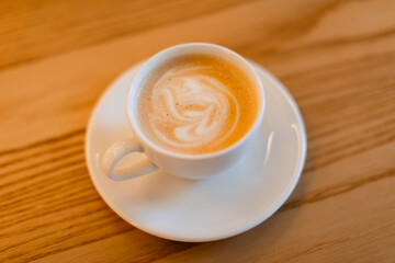 A classic cappuccino with delicate latte art served in a white ceramic cup on a matching saucer with a silver spoon. The warm wooden table adds a cozy coffee shop ambiance.