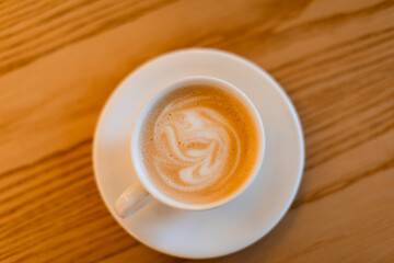 A classic cappuccino with delicate latte art served in a white ceramic cup on a matching saucer with a silver spoon. The warm wooden table adds a cozy coffee shop ambiance.