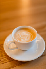 A classic cappuccino with delicate latte art served in a white ceramic cup on a matching saucer with a silver spoon. The warm wooden table adds a cozy coffee shop ambiance.