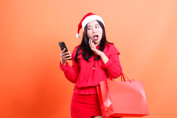 portrait of women wearing Christmas blouse, Santa hat, shocked looking at cellphone, dumbfounded hands holding device, carrying red bag of Christmas gifts, isolated on orange background
