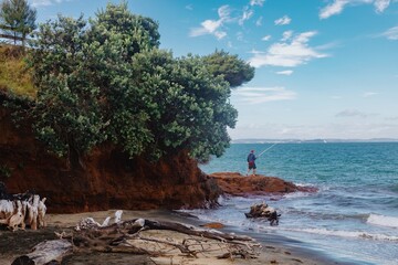 Man fishing at the beach, enjoying a tranquil day. Coastal scenery with driftwood and waves. POUTO, TE KOPURU, NORTHLAND, NEW ZEALAND. 10 Febrary 2023
