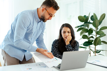 A worried man in glasses and a blue shirt and a woman in a shirt sitting at a desk, both looking at...