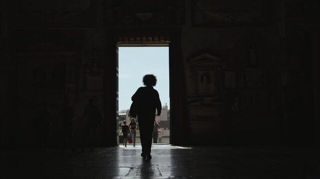 Woman is silhouetted walking at exit in the entryway of a grand Basilica di Santa Maria in Aracoeli stepping from the interior darkness into the bright sunlight