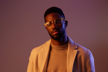 Obraz premium Stylish black man in glasses wearing a beige sweater and coat, posing with a serious expression against a purple background