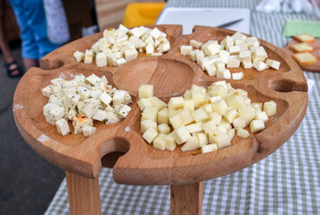 Different types of cheese are displayed on a wooden platter at an outdoor market, offering a gourmet tasting experience. The setup invites visitors to sample and enjoy assorted flavors
