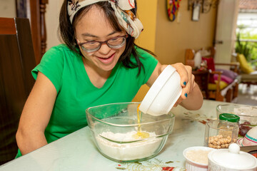 A young woman with Down syndrome, wearing glasses, empties an egg into a container full of flour. She wears an apron, concentrates on her culinary activity and enjoys the Trisomy 21 process.