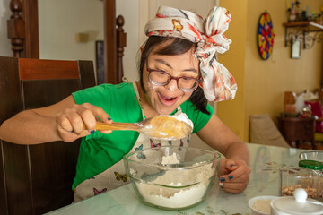 Young woman with Down syndrome, wearing glasses, enjoys mixing flour in a bowl. She wears an apron,...