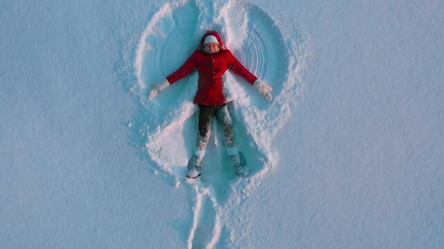 Aerial drone view girl in red winter coat making snow angel. Girl waves arms and legs lying in snow. High quality 4k footage