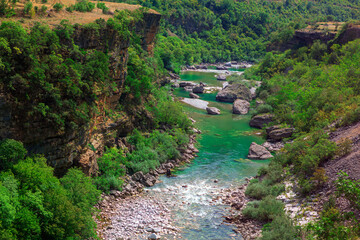 Fototapeta premium Stunning Moraca River winding through Montenegro's lush, green canyon. Rocky cliffs frame clear greenish waters dotted with large rocks and boulders. Dense vegetation adds natural allure of landscape