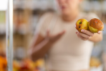 In store, blurred frustrated girl holds rotten fruits in hands, close-up. She accidentally took spoiled fruits from showcase box, fastidiously examines unsuitable goods.