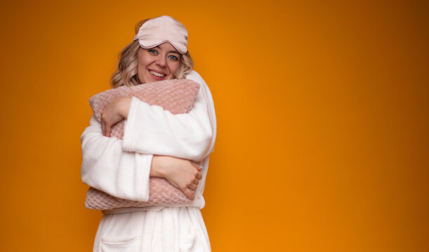 Woman in white robe and pink sleep mask, smiling and hugging soft pink pillow, set against a vibrant orange background. The image exudes warmth, comfort, and positivity, perfect for cozy home themes