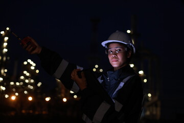 A female chemical worker in safety gear uses a digital tablet and walkie-talkie at a brightly lit refinery at night