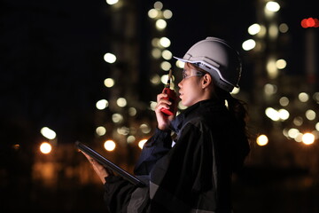 A female chemical worker in safety gear uses a digital tablet and walkie-talkie at a brightly lit refinery at night