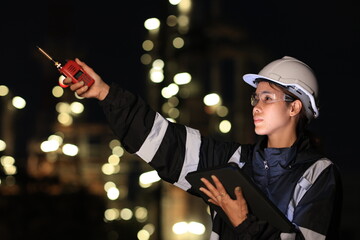 A female chemical worker in safety gear uses a digital tablet and walkie-talkie at a brightly lit refinery at night