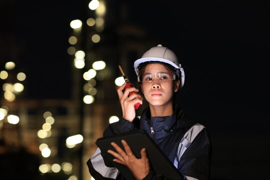 A female chemical  worker in safety gear uses a digital  tablet and walkie-talkie at a brightly lit refinery at night