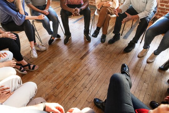 A diverse group of people sitting in a circle on wooden floor, engaged in discussion. The group includes men and women, communication. Diverse people sitting in a circle of support from community.