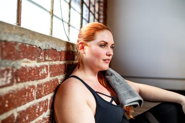 Woman resting after workout, sitting by brick wall. Fitness and relaxation. Red hair plus size woman in athletic build. Casual gym setting, towel on woman's shoulder. Post-exercise calm.