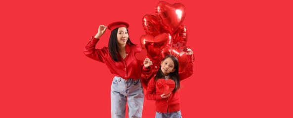 Young Asian woman and her little daughter holding gift for Valentine's day with heart-shaped balloons on red background