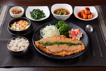 Pork cutlet and salad and parsley are placed on a black plate, and rice, soybean paste stew, vegetables, and kimchi are on the side.