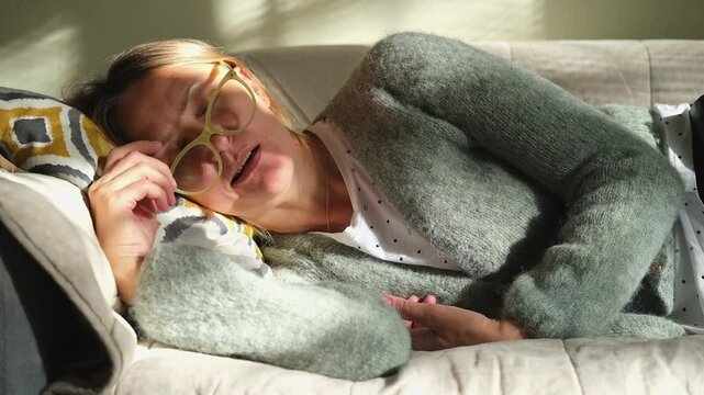 Mature woman is lying on couch at home with closed eyes. Middle aged woman is tired and exhausted. Health problems, suffers migraine, high blood pressure, aging process or menopause concept. 