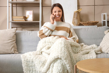 Young woman with warm plaid talking by mobile phone at home on winter day