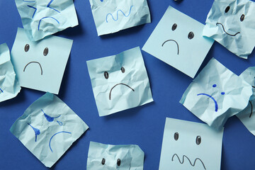 Paper sheets with drawn sad faces on blue background, top view